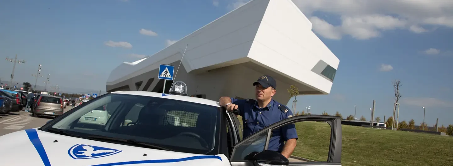 Una guardia giurata in uniforme appoggiata alla portiera aperta di un'auto di pattuglia bianca Cosmopol Spa.