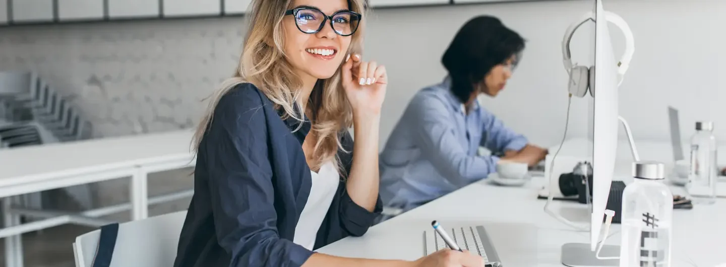 Una giovane donna sorridente con occhiali lavora a una scrivania moderna in un ufficio luminoso.