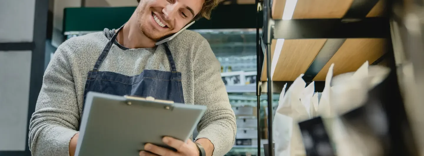 Un commerciante sorridente con grembiule prende appunti su una cartellina mentre parla al telefono nel suo negozio.
