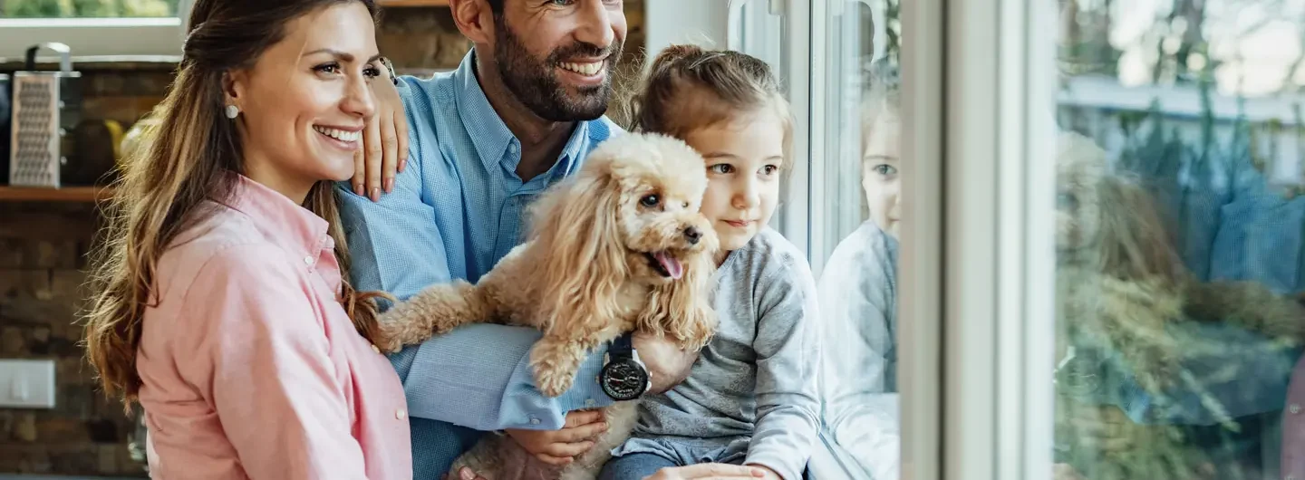 Una famiglia composta da madre, padre e figlia piccola tiene in braccio un cane mentre guarda fuori dalla finestra di casa.