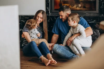 Una giovane famiglia composta da genitori e due figli piccoli seduti insieme sul pavimento davanti a un camino acceso.